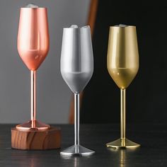 three wine glasses sitting next to each other on top of a wooden stand, one gold and the other silver