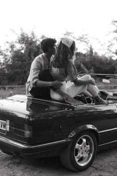 two women sitting on the hood of a convertible car in black and white, with trees in the background