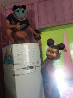 a man in mickey mouse mask sitting on top of a refrigerator next to a woman