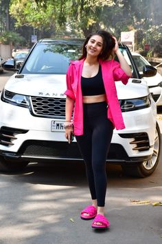 a woman in pink jacket and black top standing next to a white car with her hand on her head