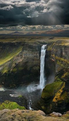 an image of a waterfall in the middle of some land with clouds above it and water below