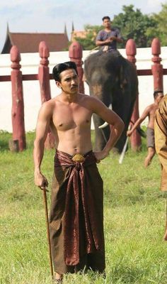 a man with no shirt standing in the grass next to an elephant and holding a stick