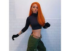 a woman with red hair is posing in front of a white wall and wearing green pants