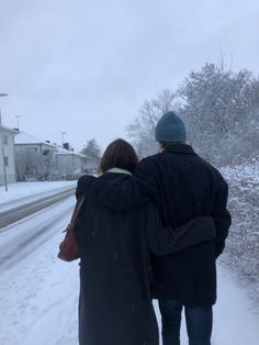 a man and woman walking down a snow covered road in the middle of winter with their arms around each other