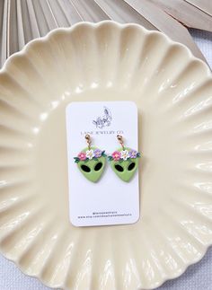 a pair of earrings with an alien head and flowers on the ears are sitting on a plate