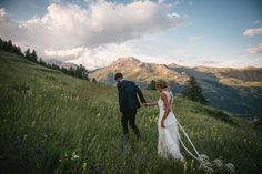 a bride and groom holding hands walking through tall grass with mountains in the back ground