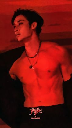 a man with no shirt on standing in front of a red wall and wearing a necklace