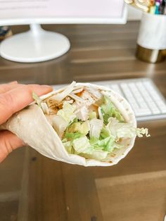 a person is holding a burrito with lettuce and chicken on it in front of a computer desk