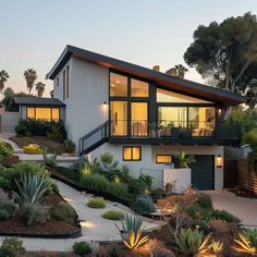 a modern house is lit up at night in the evening with plants and trees surrounding it