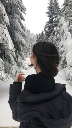 a woman standing in front of snow covered trees and looking at the ground with her back to the camera