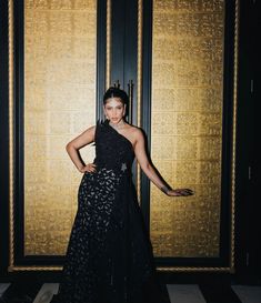 a woman standing in front of a gold wall wearing a black dress and holding her arms out