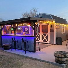 an outdoor bar is lit up with colorful lights and barrels in the foreground, next to a shed