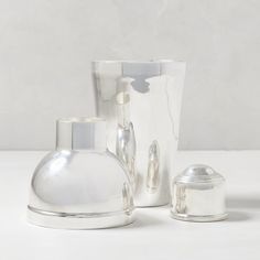 two silver vases sitting next to each other on a white counter top with a cup in the background