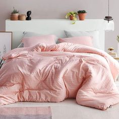 a bed with pink comforter and pillows in a white room next to potted plants