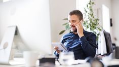 a man sitting in front of a computer talking on a cell phone and holding a clipboard