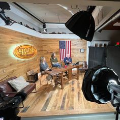 two people sitting at a table in front of an american flag on the wall behind them