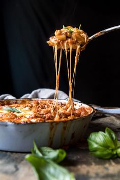 a spoon full of spaghetti being lifted from a casserole dish with basil leaves