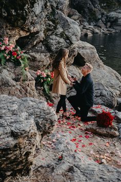 a man kneeling down next to a woman on top of a rocky cliff near water