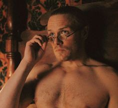 a shirtless man with glasses on sitting in a chair holding a cell phone to his ear