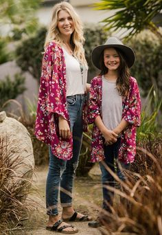 two women standing next to each other in front of trees and bushes, one wearing a pink floral kimono