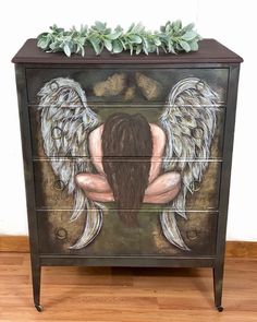 an artistic painted dresser with angel wings on the front and sides, sitting on top of a hard wood floor