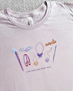 For all the nostalgic girlies who miss certain movies from the 2000s ✨ This tee features the words "Live your own fairy tale" below eight magical icons, each hand-drawn in colored pencil: a wand of light, a tiara made from a magic crystal, dancing slippers, the Sugar Plum Princess locket, a necklace that will grant you fairy wings, a royal crown, a stone from the Diamond Castle, and an enchanted paintbrush. This shirt is also available without the "Live your own fairy tale" tagline. SIZING AND COLOR OPTIONS: Please see the images in the gallery. Please note, due to differences in how screens display color, the actual color of the product may vary from what is shown. The Lavender Dust color is pale gray with a tinge of lavender, a perfect backdrop for these delicate illustrations. Artwork i Movies From The 2000s, Sugar Plum Princess, Rapunzel Tattoo, Princess Disneybound, Magic Of Pegasus, Silly Shirts, Barbie 12 Dancing Princesses, Magic Crystal, 12 Dancing Princesses