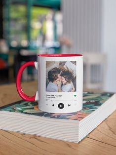 a red and white coffee mug sitting on top of a wooden table next to a book