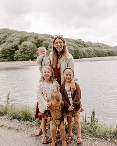 a woman standing next to two children near a body of water with trees in the background