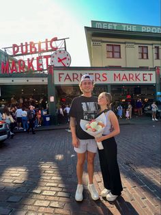 a man and woman standing in front of a farmers market with their arms around each other