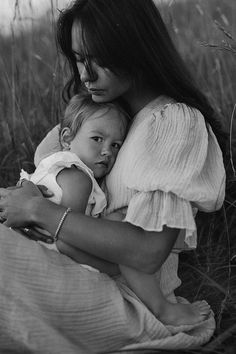 a woman holding a baby while sitting on top of a grass covered field in front of tall grass