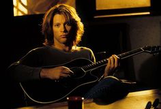 a man with long hair playing an acoustic guitar in a dimly lit room, looking at the camera