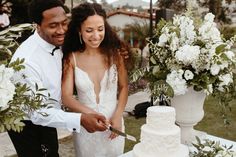 a man and woman cutting into a white wedding cake on top of a table with flowers