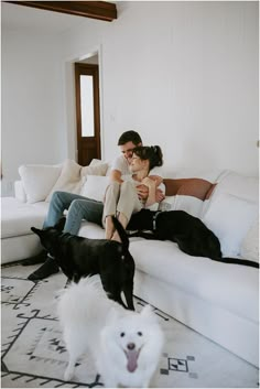 a man and woman sitting on a couch with two dogs in the living room next to them