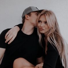 a man and woman sitting next to each other in front of a white wall with their arms around each other