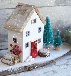 a small white house sitting on top of a piece of wood next to christmas trees