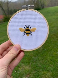 a hand holding up a small cross stitch bee in front of a green field with trees