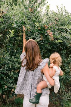 a woman holding a toddler in her arms while standing next to bushes and trees