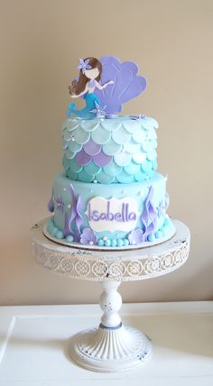 there is a blue and purple cake with a little mermaid on the top, sitting on a white pedestal