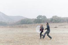 two people running across a field in the rain with an umbrella over their heads,