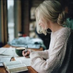 a woman sitting at a table with a book and pen in her hand while writing