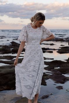 a woman in a white dress standing on rocks near the ocean with her hands behind her back