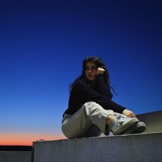 a woman sitting on top of a cement wall next to a blue and pink sky