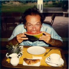 a man sitting at a table eating a piece of watermelon with his mouth open