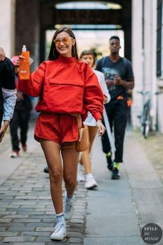 a woman in red is walking down the street while holding a bottle and wearing white sneakers