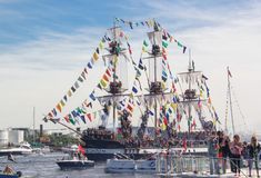 several people are standing on the dock looking at a large boat with flags flying from it