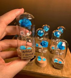 a hand holding a tiny blue rose in a glass bottle with other small ones behind it