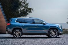 a blue suv parked on gravel next to the ocean with trees and water in the background