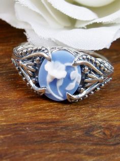 Blue Cameo Orchid RingSmall Oval Gothic Design#D85 Inspired by Gothic designs, this stunning antique reproduction ring is crafted in sterling silver. This gorgeous ring is set with a flawless oval simulated cameo. The outline of an orchid on a soft blue background is highlighted in white. The stunning cameo is 11mm x 9mm. The band is marked 925 for sterling silver. Notice the beautiful silver filigree swirl setting and flared band. The bold style travels around caressing the central focus while Victorian Silver Carved Rings, Victorian Carved Silver Rings, Victorian Style Carved Silver Rings, Vintage Blue Carved Jewelry, Vintage Carved Blue Jewelry, Victorian Sterling Silver Rings With Cabochon, Victorian Silver Cameo Rings, Vintage Sterling Silver Cameo Ring, Vintage Sterling Silver Flower Ring With Intricate Design