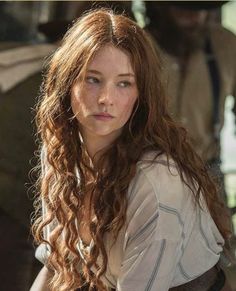 a woman with long red hair sitting on a bench next to a man in the background