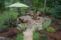 a stone patio with chairs and an umbrella in the middle is surrounded by trees, shrubs, and flowers